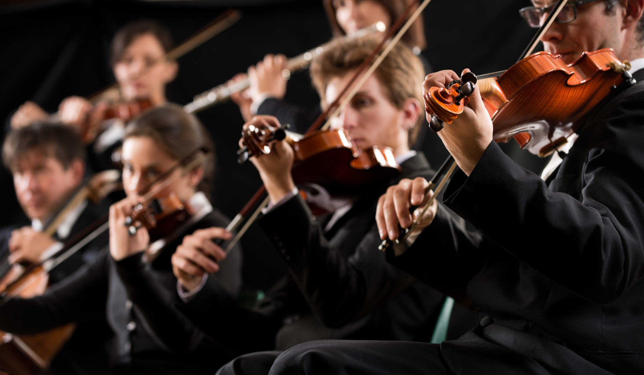 Symphony orchestra first violin section performing on dark background.